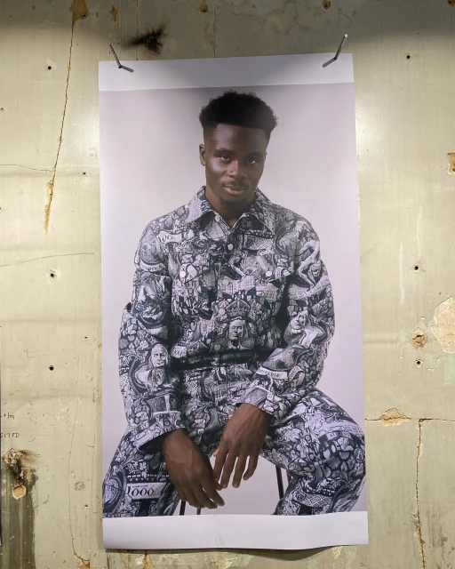 Portrait of a Black male model wearing a black and white patterned jacket and pants, sitting in front of a cream wall, poster.