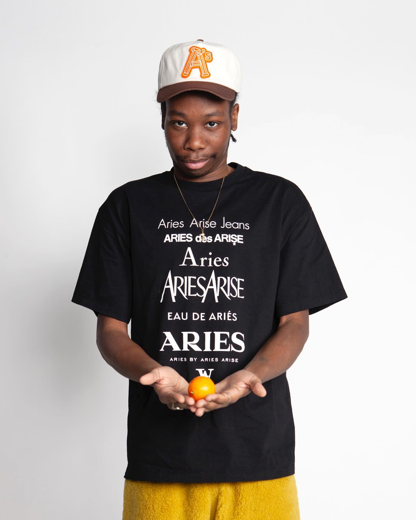 An adult male wearing a baseball cap and graphic t-shirt holds an orange in a studio shot on a white backdrop, demonstrating streetwear fashion.