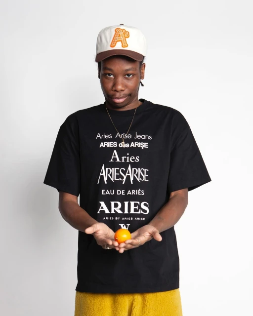 An adult male wearing a baseball cap and graphic t-shirt holds an orange in a studio shot on a white backdrop, demonstrating streetwear fashion.