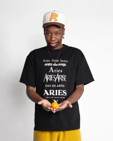 An adult male wearing a baseball cap and graphic t-shirt holds an orange in a studio shot on a white backdrop, demonstrating streetwear fashion.