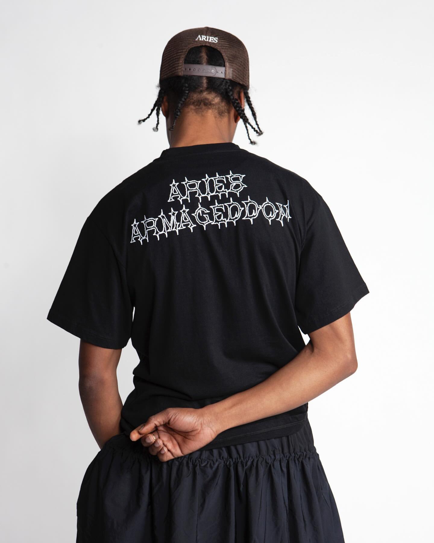 An adult male is seen from the back wearing a black t-shirt with white writing and a brown baseball cap against a white backdrop.