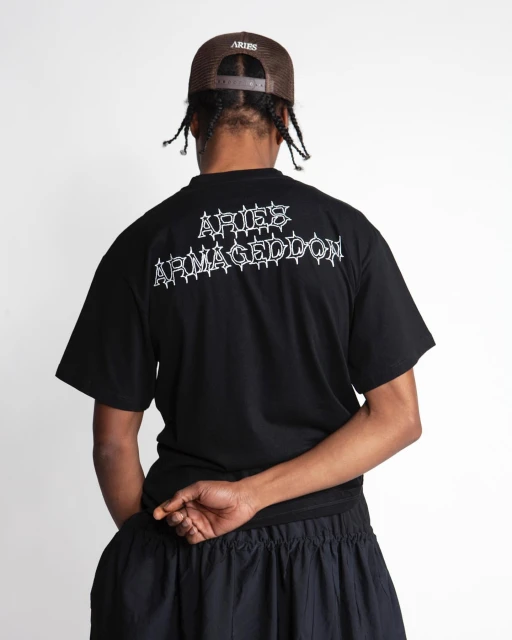 An adult male is seen from the back wearing a black t-shirt with white writing and a brown baseball cap against a white backdrop.