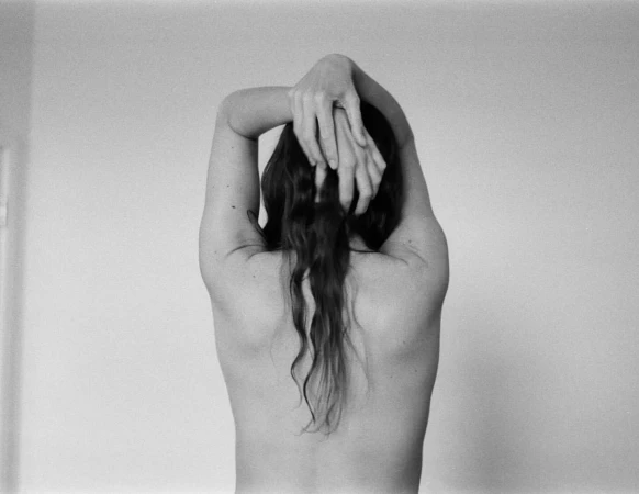 Back view of a slender adult female with dark hair, hands clasped behind her head, posing against a neutral, light-grey background.