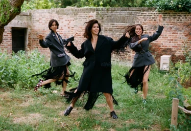 Three women dancing outdoors in an overgrown garden wearing dark oversized blazers with fringe detailing, photographed by Clare Shilland.