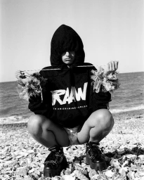 Black and white portrait of a person crouching on a pebble beach wearing a hooded sweatshirt and platform boots.