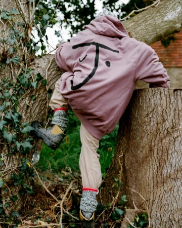 Person in mauve hoodie with a black graphic on back climbing a large tree in a wooded setting, photographed by Clare Shilland