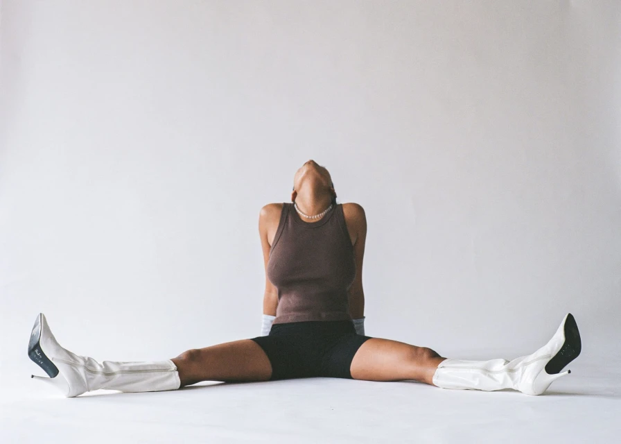 Woman poses with legs spread wearing shorts, top, pearl necklace and boots against white studio backdrop, in a fashion pose.