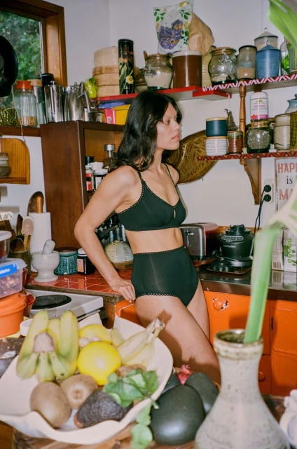 Woman in a black bra and high-waisted bottoms poses in a kitchen with a bowl of fruit, near white walls and wooden cabinets.