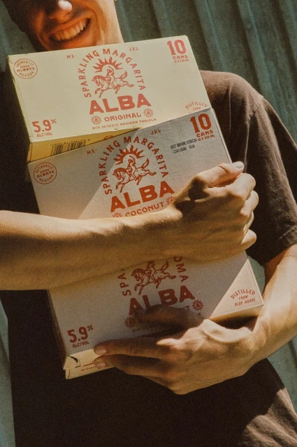 Smiling man holds stacked ALBA sparkling margarita 10-can boxes outdoors in soft, warm lighting, showcasing a casual, beverage-focused scene.