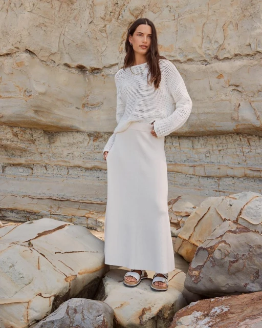 A woman in a white knit sweater and cream maxi skirt stands outdoors on rocks with a rock wall behind, fashion shoot.