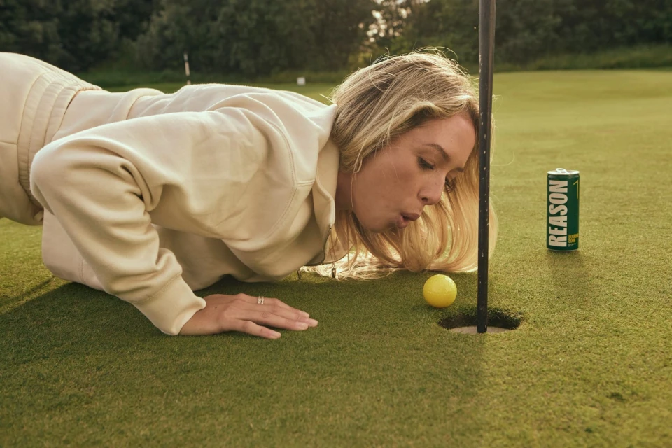 Blond woman in a beige tracksuit blowing a yellow golf ball toward the hole on a golf course; a can of 'Reason' is on the green.