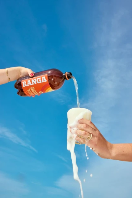 Beverage commercial with a brown soda bottle pouring into a clear, overflowing glass held by women's hands against a blue sky.