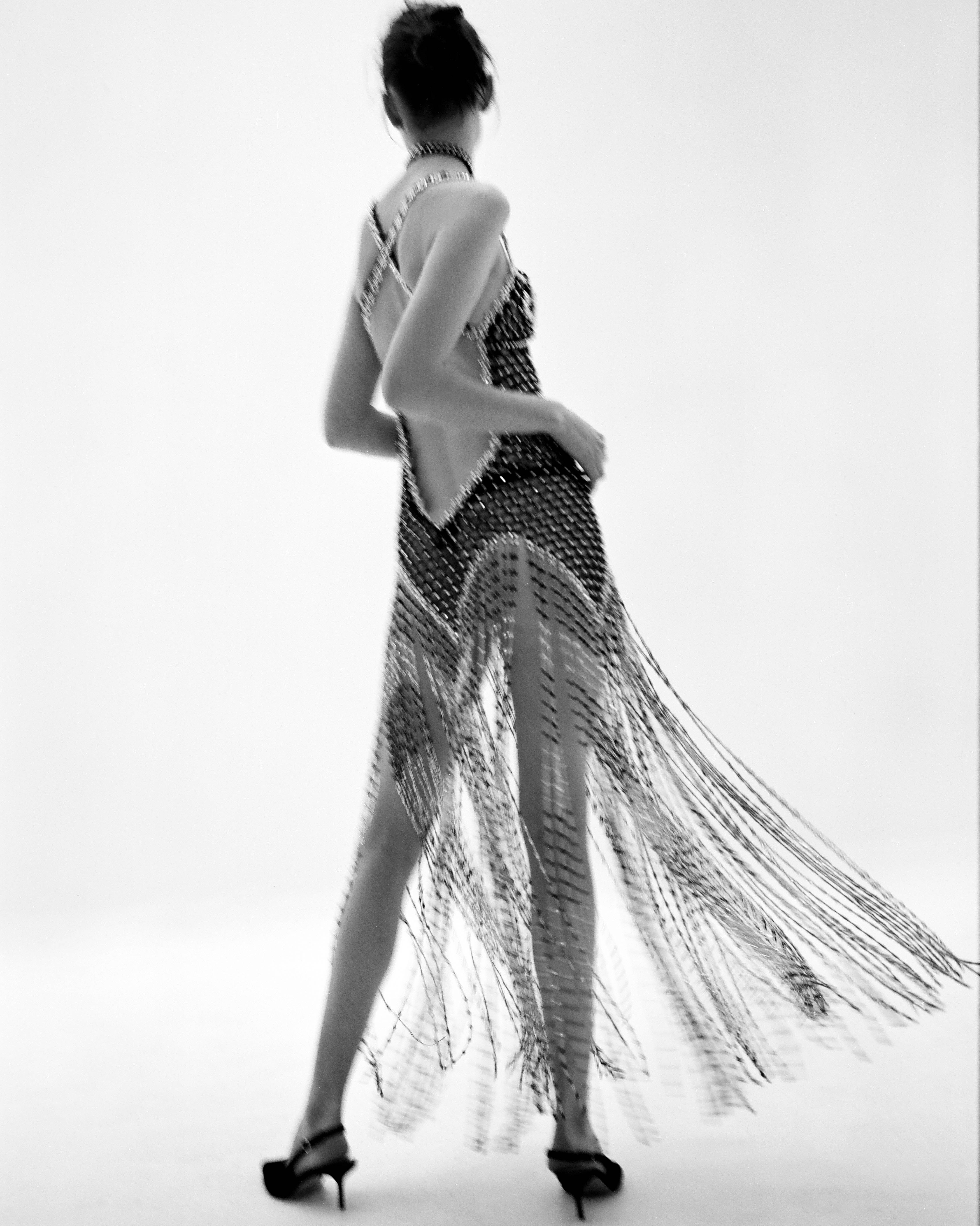 Back view of a slender adult woman with a blurred motion effect, wearing a fringed sleeveless dress and heels in a white setting.