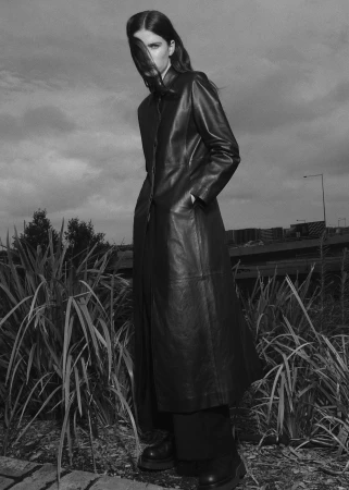 Monochrome fashion portrait of a woman wearing a long leather coat standing among tall grass outdoors.