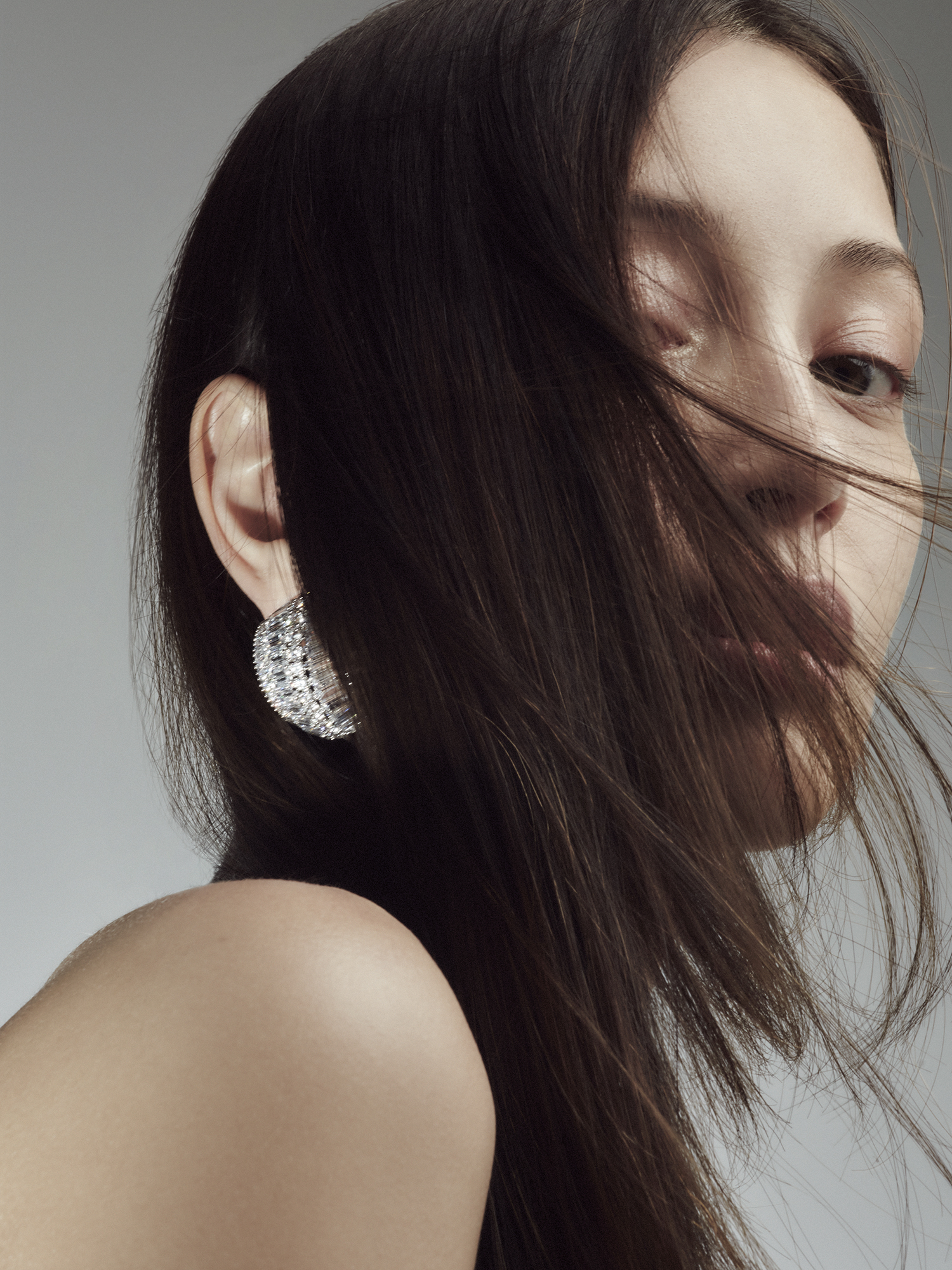 Close-up of a woman with dark hair, wearing a glittering earring, posing against a soft grey background in a beauty or fashion context.