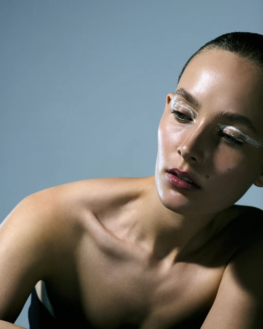 Close-up of a woman with fair skin and exaggerated white makeup on her lashes and brows against a solid blue background.