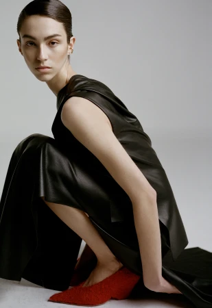 Model in black sleeveless top and trousers crouching down wearing red shoes against a grey background
