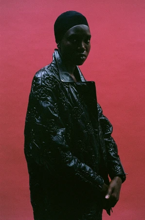 Black woman wearing a dark embossed leather coat and matching cap against a red background by Sheree Porter