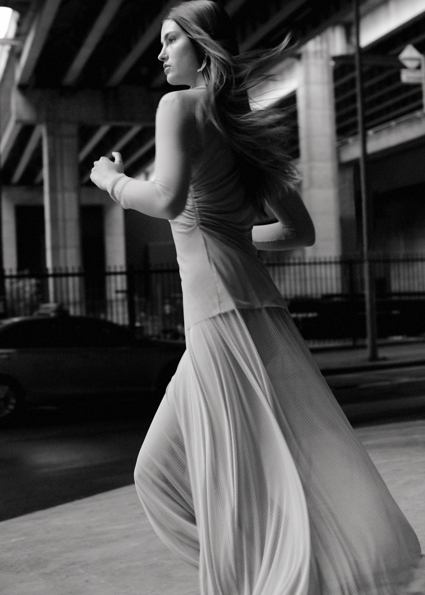 Monochrome fashion shot of a woman in a flowing dress running beneath an overpass, captured in a medium shot with diffused lighting.