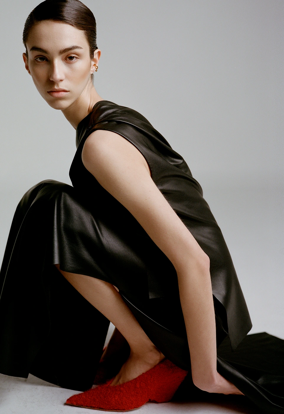 Model in black sleeveless top and trousers crouching down wearing red shoes against a grey background