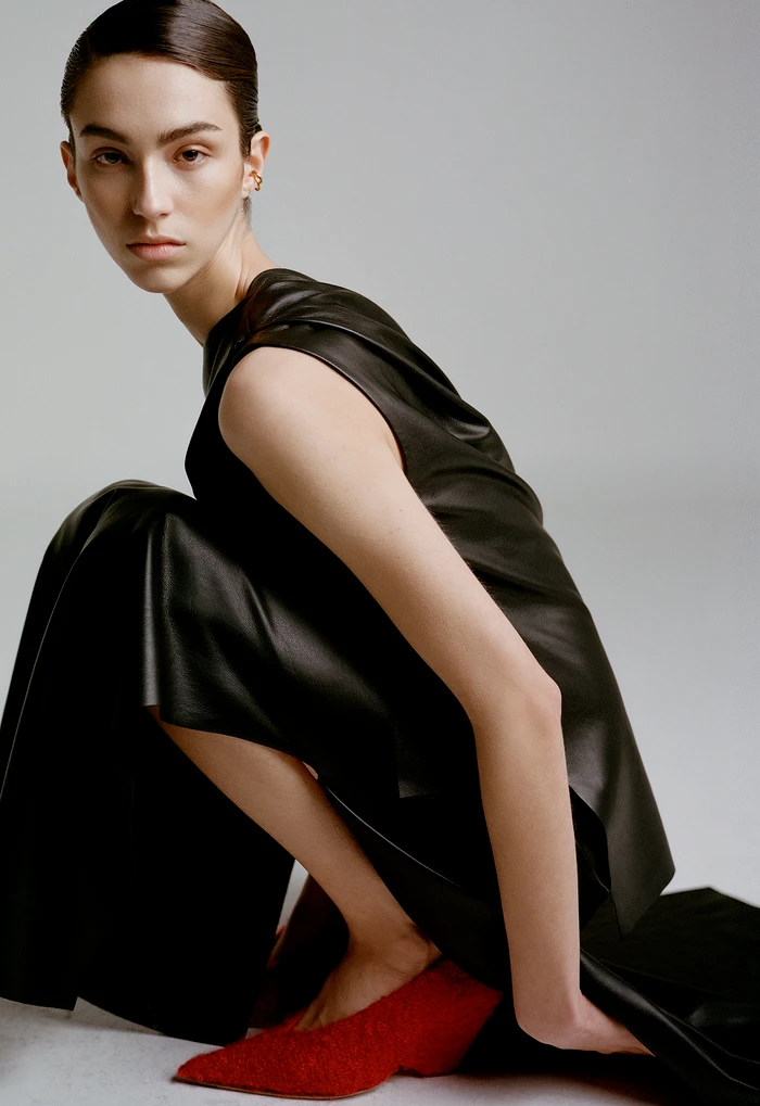 Model in black sleeveless top and trousers crouching down wearing red shoes against a grey background