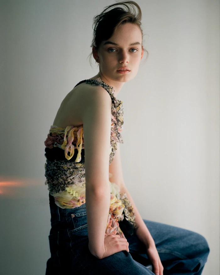 A woman in a textured multi-color sleeveless top and blue jeans sits sideways against a pale grey background.