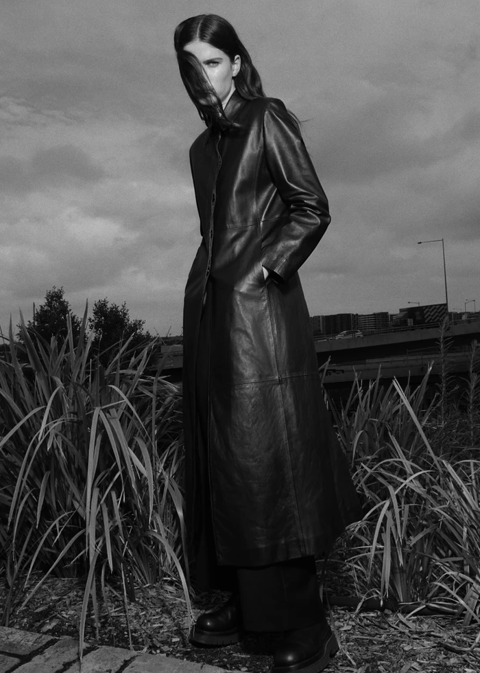 Monochrome fashion portrait of a woman wearing a long leather coat standing among tall grass outdoors.