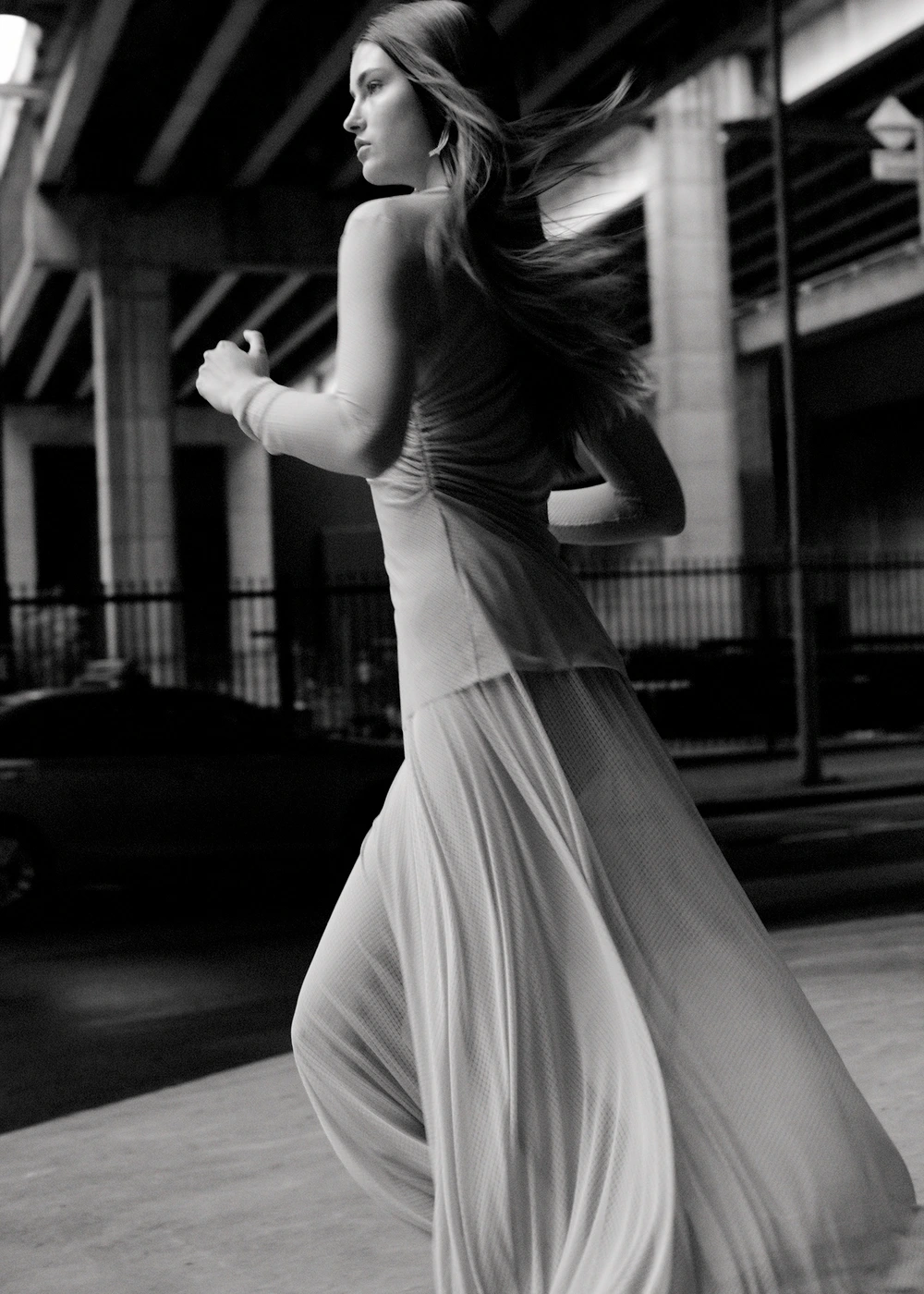 Monochrome action shot of a woman with long hair running outdoors near an elevated roadway structure, wearing a pleated skirt.