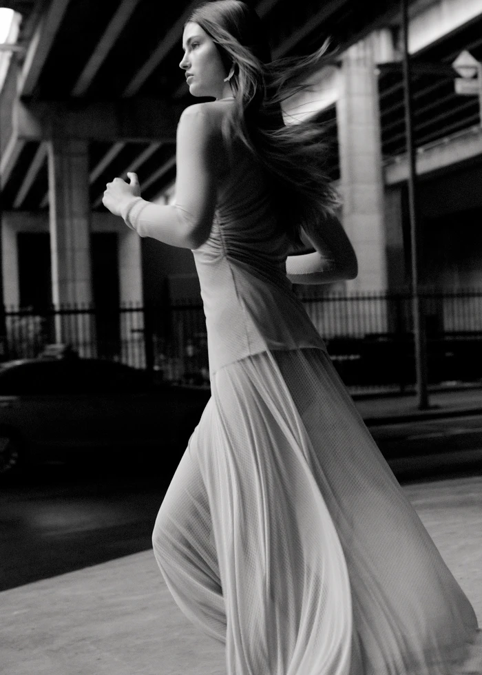 Monochrome action shot of a woman with long hair running outdoors near an elevated roadway structure, wearing a pleated skirt.