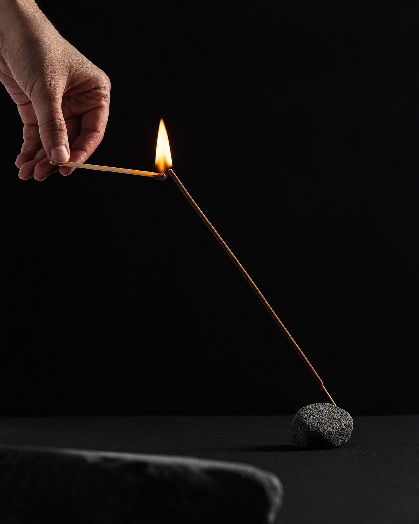 Hand ignites incense stick with match in dark setting. Incense stick in a stone holder, photographed indoors with a black background.