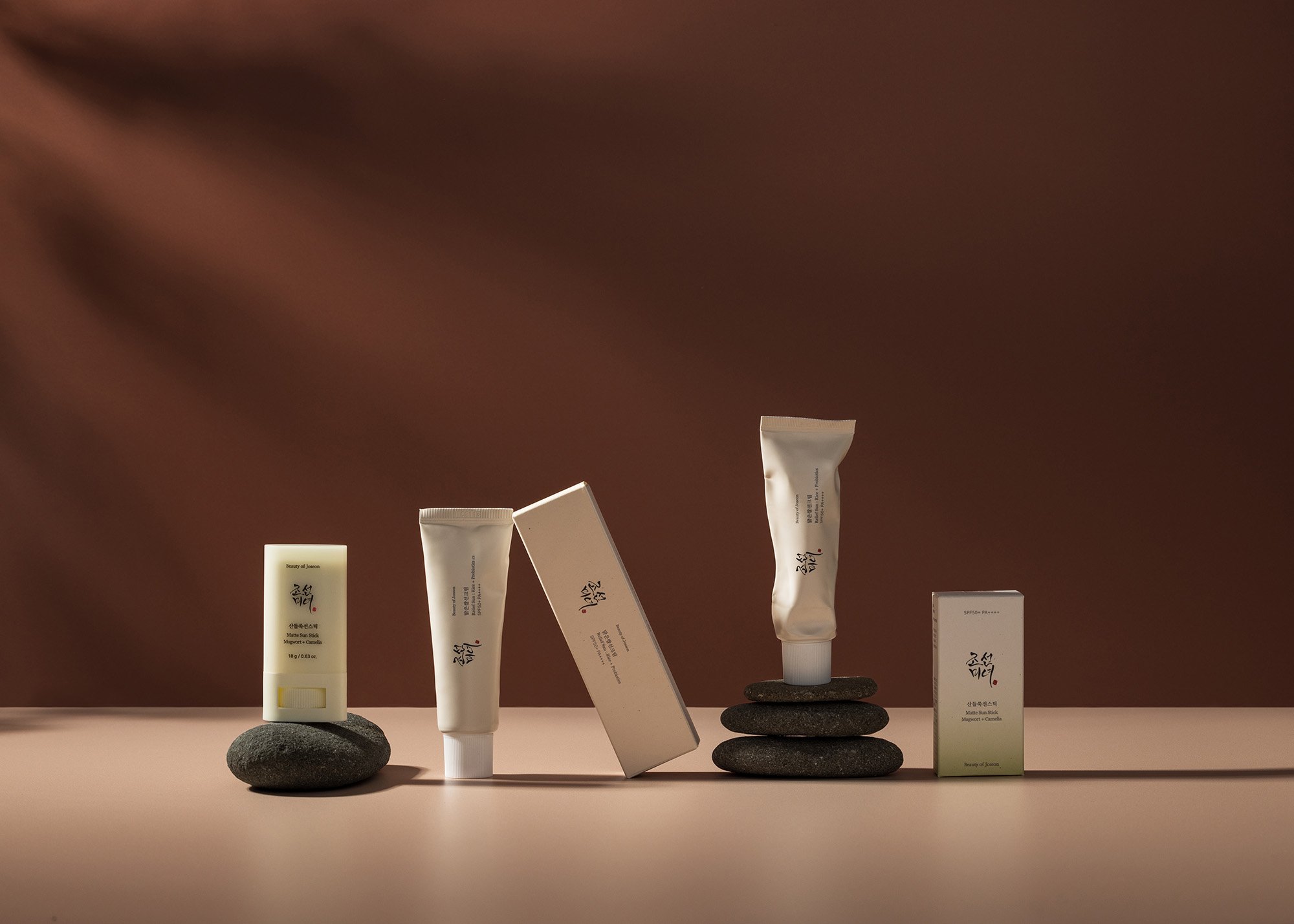 Five cosmetic containers arranged on pebbles against a brown backdrop with soft lighting, conveying a minimalist aesthetic.