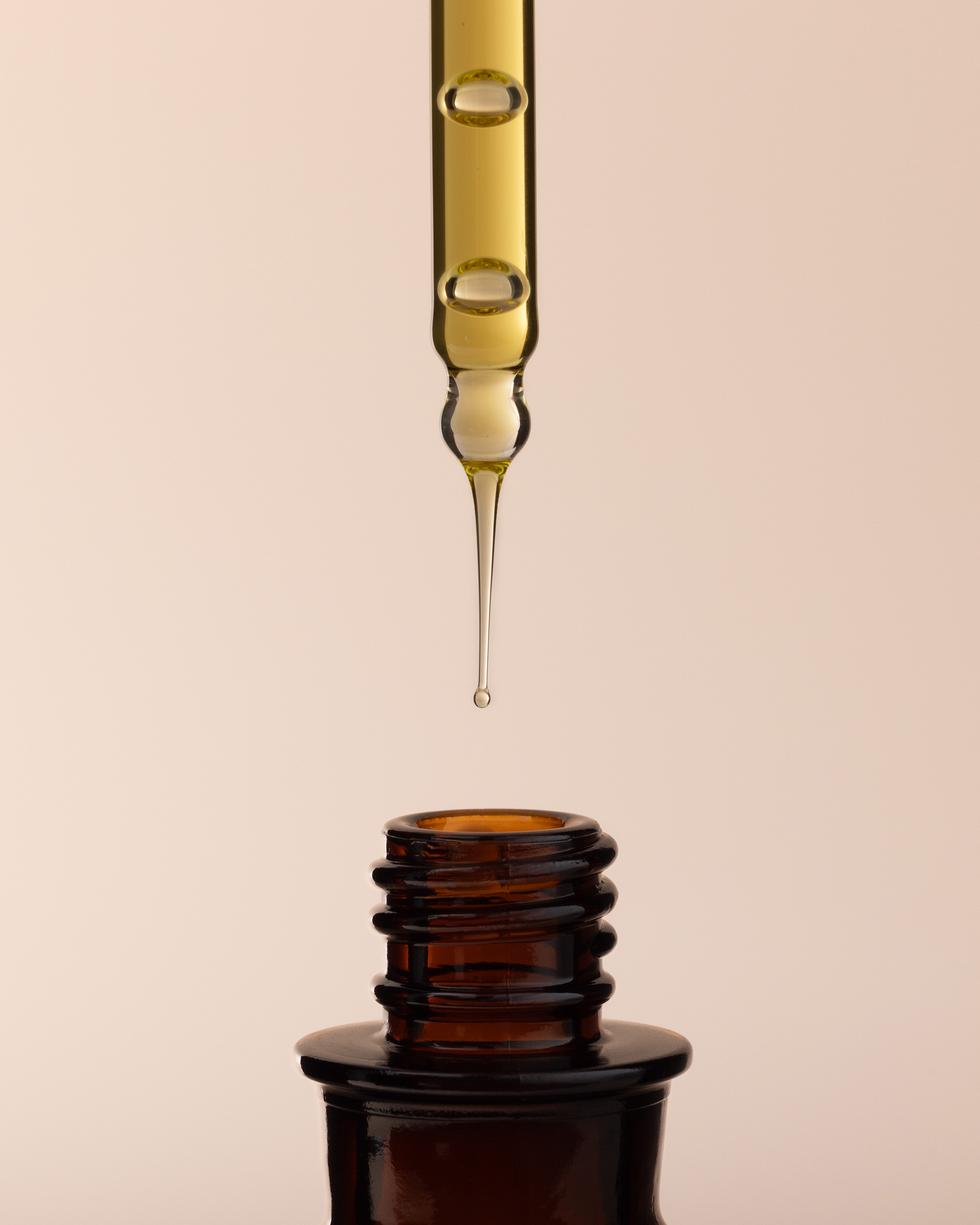 Close-up of amber-colored dropper dispensing liquid into a brown glass bottle, studio shot on a soft, neutral background.