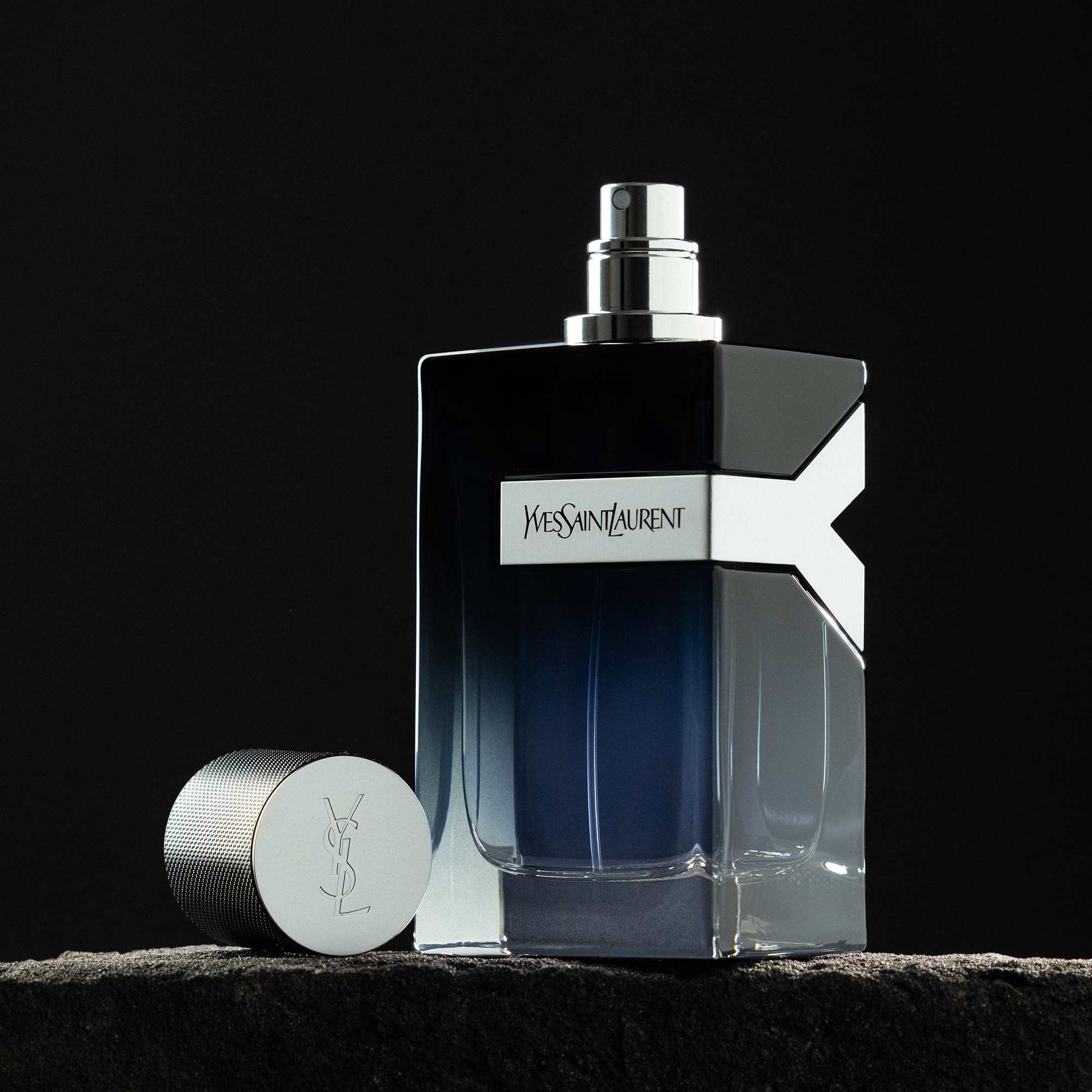 Studio shot of YSL Y Le Parfum fragrance bottle with silver cap on dark stone against a black background. Elegant and modern design.