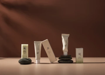 Skincare product tubes and boxes arranged on dark stones against a warm brown backdrop with natural light shadows