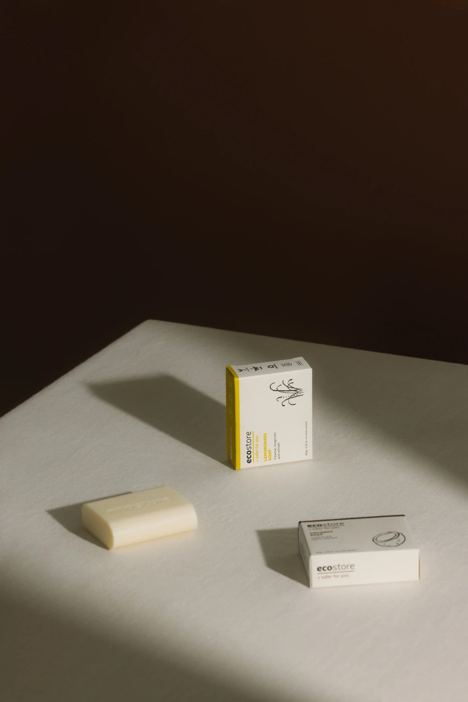 Product shot of a bar of soap and two product boxes on a white surface with warm diagonal shadows cast across the scene