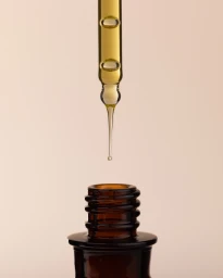 Glass dropper dispensing a golden serum drop above an amber bottle against a neutral beige background.