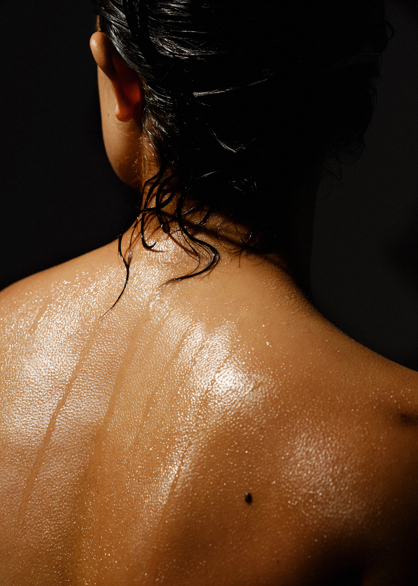 Close-up shot of a woman's back showcasing her wet hair and glistening skin against a dark background, emphasizing texture and contour.