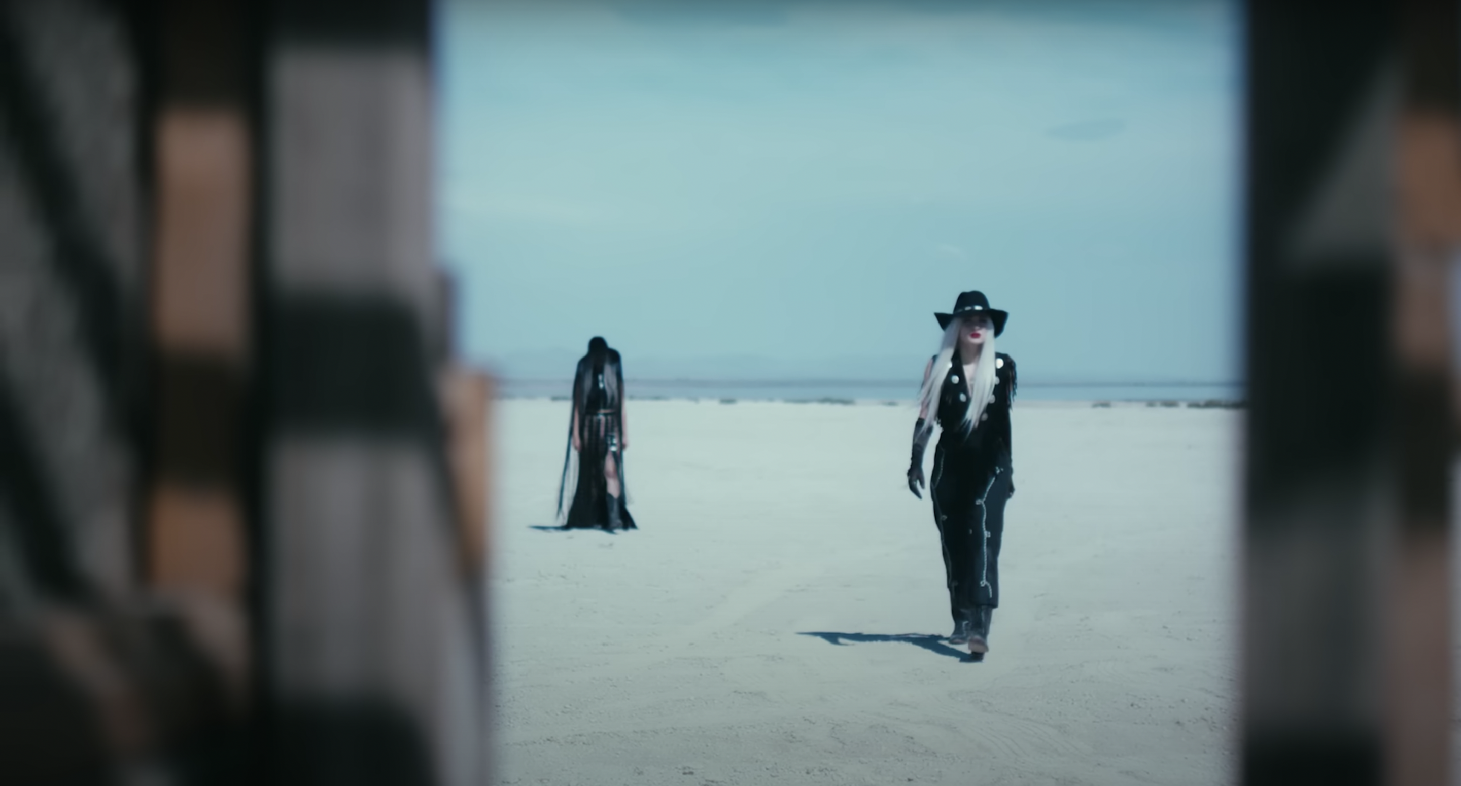 Two women in distinct black outfits walk on a desolate beach under a pale sky, framed by blurred wood for a cinematic effect.