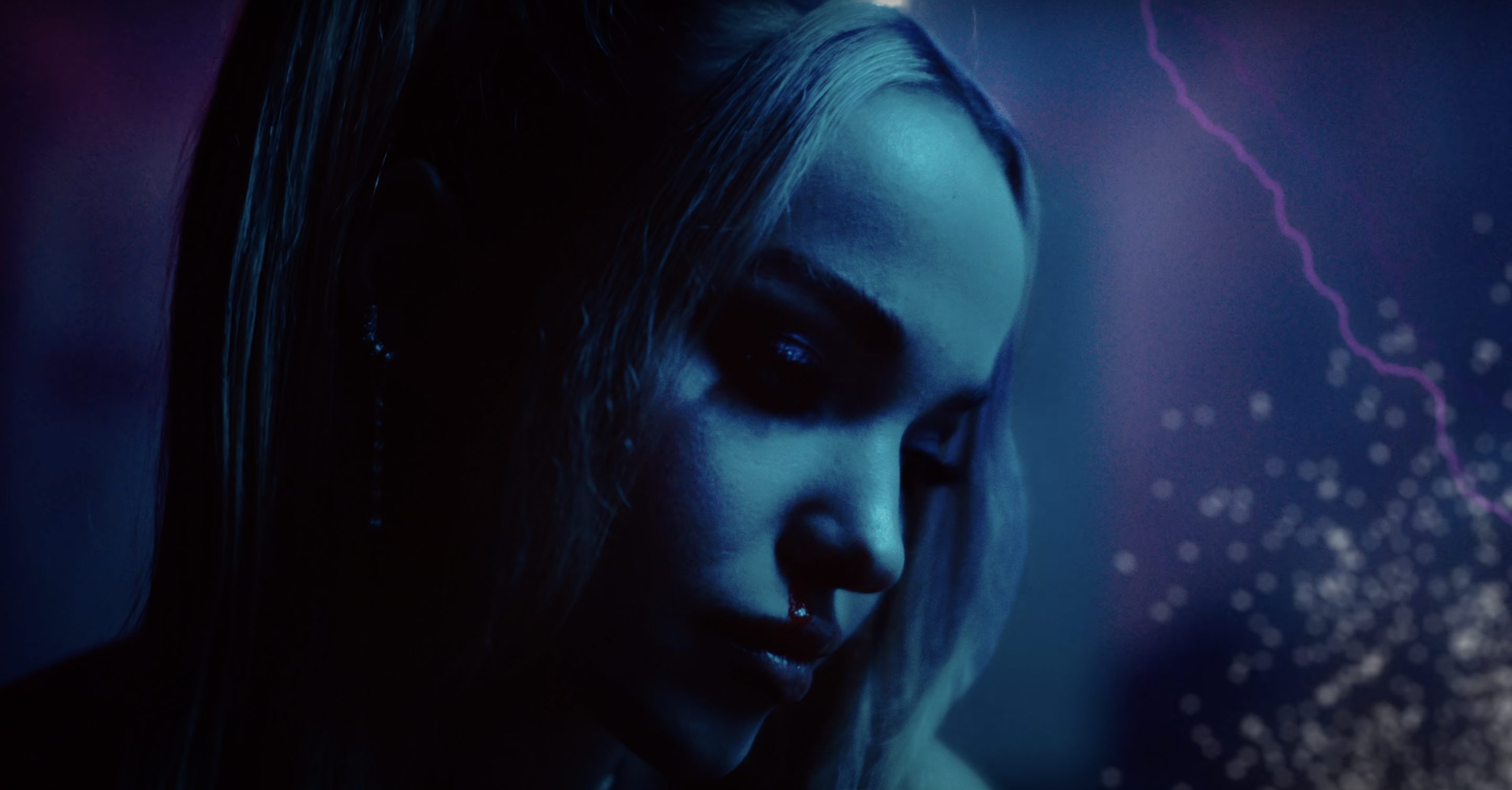 Close-up of a young adult female with light blonde hair, blue eyes, and fair skin in a dark indoor setting with a blue color cast.