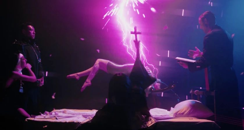 Woman levitates in a dimly lit scene with neon lighting, surrounded by a magician, assistants, a cross, and electrical sparks.