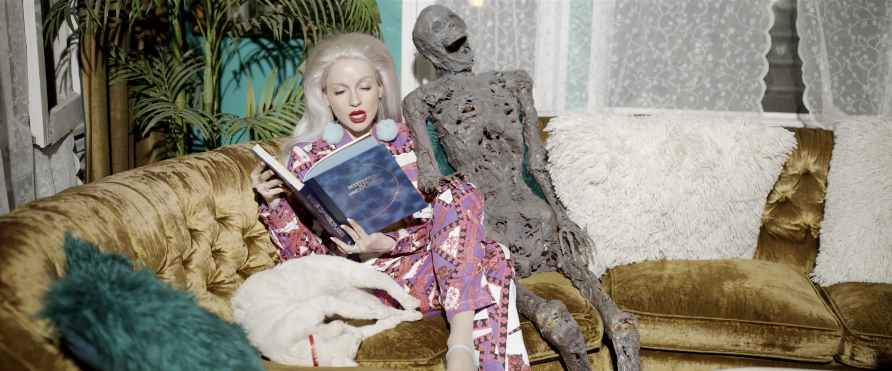 Woman in patterned pajamas reads a book on a gold couch with a white cat and a sculpture of a human figure, indoor setting with plants.
