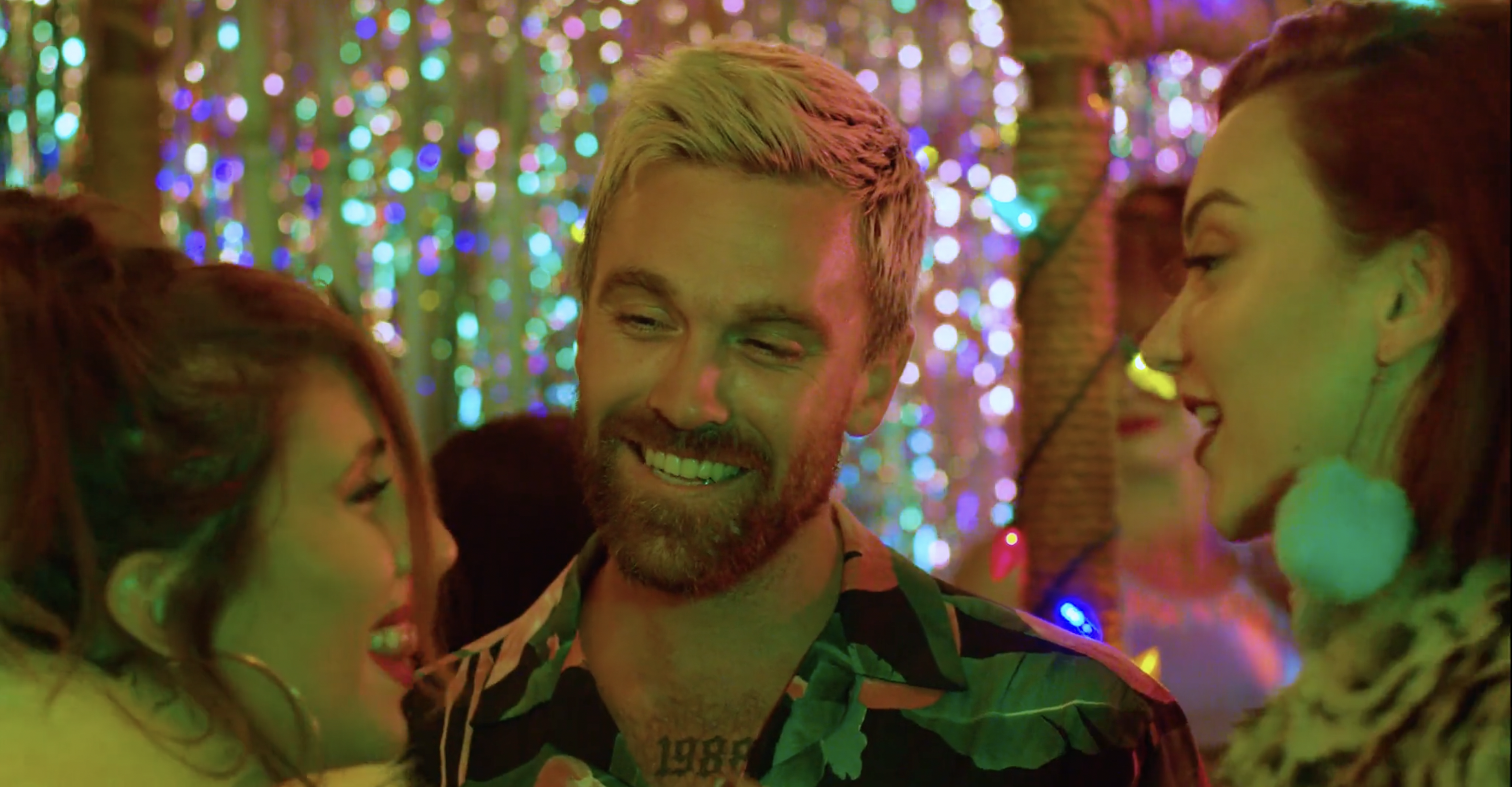 Close-up of a smiling man with two women in a brightly lit indoor setting with bokeh lights and a tropical shirt, possibly at a party.