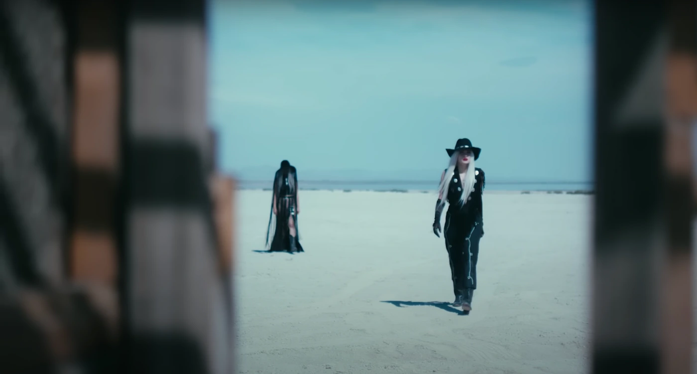 Two women in dark outfits walking across a vast open salt flat under an overcast sky framed by a doorway