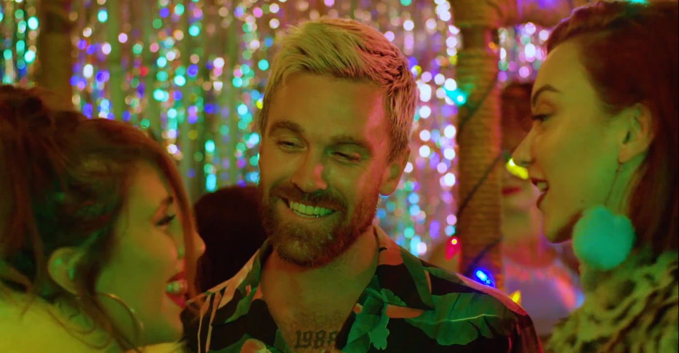 Man with blonde-tipped hair and a patterned shirt smiling surrounded by women in a colorful illuminated venue directed by Felice Vale