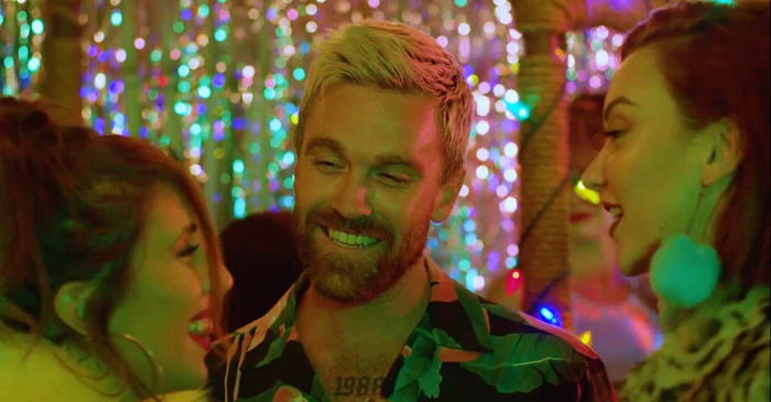 Man with blonde-tipped hair and a patterned shirt smiling surrounded by women in a colorful illuminated venue directed by Felice Vale
