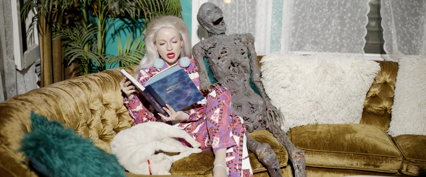 Blonde woman in patterned pyjamas seated on a gold velvet sofa reading a book next to a skeletal prop