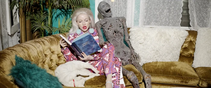 Blonde woman in patterned pyjamas seated on a gold velvet sofa reading a book next to a skeletal prop