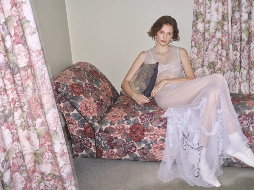 Woman poses on floral chaise lounge in sheer dress, white boots, and drop earrings with floral curtains in soft, diffused light.