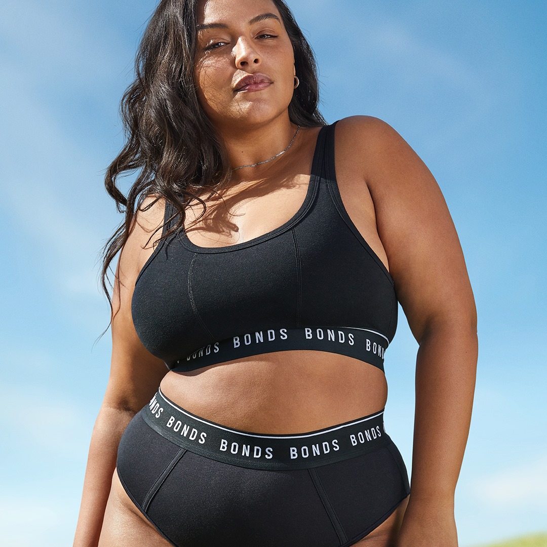 Woman in black Bonds underwear and sports bra poses outdoors against a light blue sky. Close-up shot of the model.