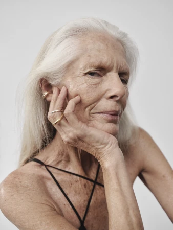 Elegant portrait of a senior woman with long gray hair. She is resting her face on her hand, posing in a studio with a plain white backdrop.